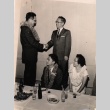 Two men standing shaking hands, man and woman seated at dinner table (ddr-njpa-4-42)