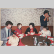 Tomoye (Nozawa) Takahashi sitting at a table with two children and a man, man with camera standing behind (ddr-densho-426-2221)