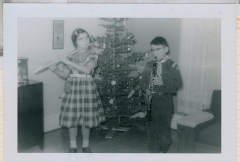 Mary Alice and David playing wind instruments in front of Christmas tree (ddr-densho-462-208)
