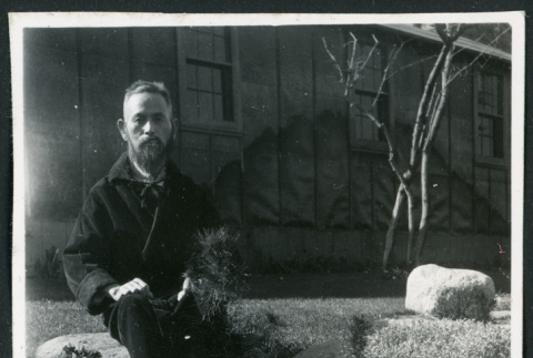 Photograph of a man in a robe sitting on a rock wall in front of the Manzanar hospital (ddr-csujad-47-245)