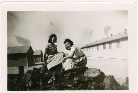 Two women sitting on pile of rocks, barracks in background (ddr-densho-329-1360)