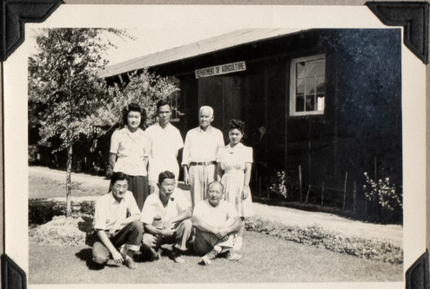 Seven people outside barrack building with sign, Department of Agriculture (ddr-densho-587-18)