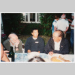 Tom Ikeda sitting in between two older men at outdoor cookout (ddr-densho-506-148)