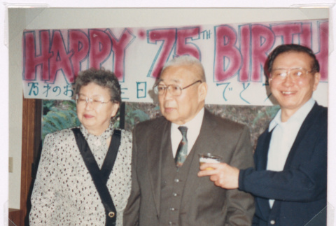 Isoshimas and brother-in-law in front of 75th birthday banner (ddr-densho-477-638)