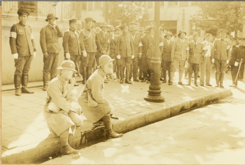 Men wearing gas masks sitting on a curb near a group of military officers (ddr-njpa-13-1180)