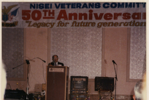 Man standing at podium under banner for 50th Anniversary Nisei Veterans Comm[...] (ddr-densho-466-552)