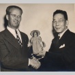 Two men shaking hands and posing with a doll (ddr-njpa-4-59)