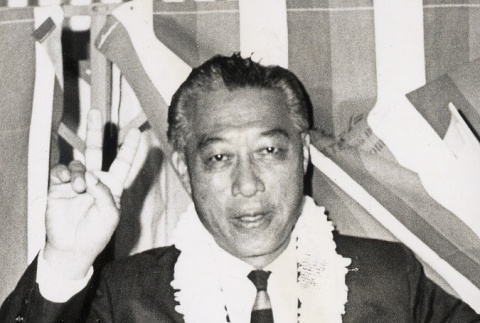 Man wearing lei, making victory sign in front of flag (ddr-njpa-2-304)