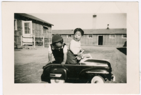 Two children in toy car with barracks in background (ddr-densho-329-1358)