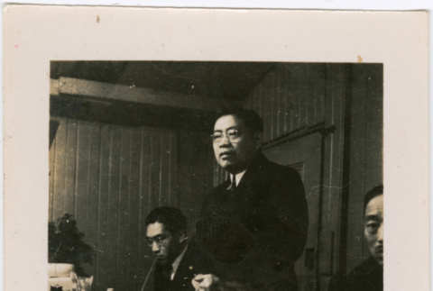 Man standing by table with food, two men seated on either side (ddr-densho-426-1933)