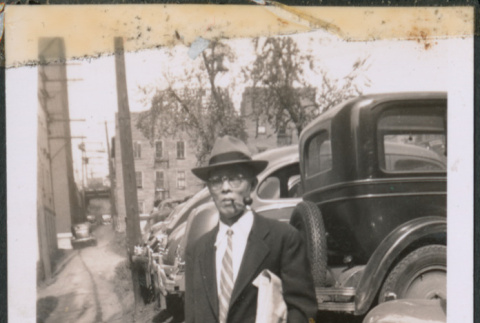Man with glasses carrying a bag standing by row of cars (ddr-densho-483-992)