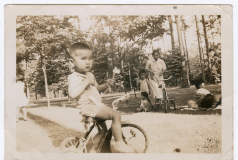 Photo of a child riding a tricycle with another child and a woman in the background (ddr-densho-483-743)