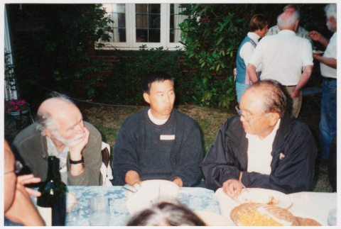 Tom Ikeda sitting in between two older men at outdoor cookout (ddr-densho-506-148)