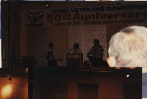 Three people on stage under banner for 50th Anniversary Nisei Veterans Committee (ddr-densho-466-555)