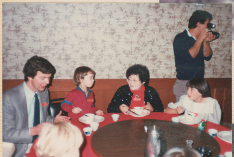 Tomoye (Nozawa) Takahashi sitting at a table with two children and a man, man with camera standing behind (ddr-densho-426-2221)