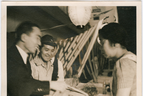 Two men handing a package wrapped in rope to woman across a table (ddr-densho-426-1871)