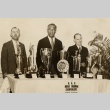Duke Kahanamoku standing behind a table of trophies with two men (ddr-njpa-2-498)