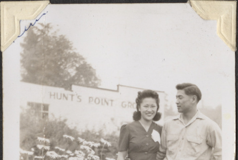 Couple standing by flowers outside Hunts Point Grocery (ddr-densho-466-904)