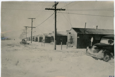 Photograph of snow covered Manzanar with a line of buildings and a car in the foreground (ddr-csujad-47-52)