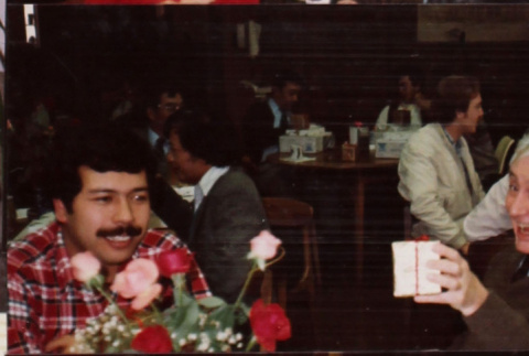Three people at a table. Man at center holding box (ddr-densho-426-1261)