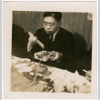 Man serving himself food from a dish with chopsticks (ddr-densho-426-1736)