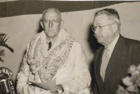 Lester Petrie wearing leis, standing with another man behind a microphone (ddr-njpa-2-812)