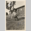 A woman kneeling next to a tree in front of barracks (ddr-densho-420-41)
