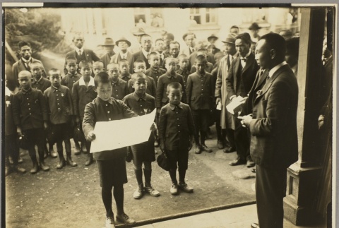 A boy reading a speech in front of men and other schoolboys (ddr-njpa-13-1189)