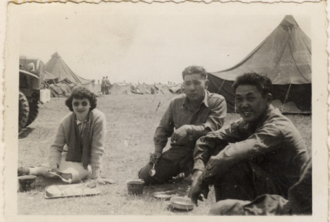 Woman and two men eating on ground near tents (ddr-densho-466-378)