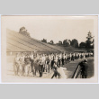 Large group of men marching on track in stadium with photographers (ddr-densho-329-1369)