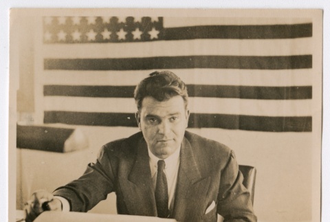 Man in suit sitting in front of the American flag (ddr-densho-223-2)