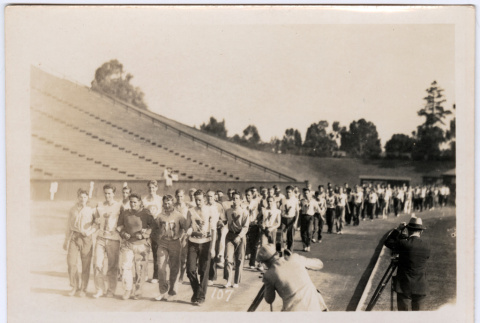 Large group of men marching on track in stadium with photographers (ddr-densho-329-1369)