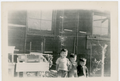 Photograph of three small boys standing in front of a bararcks with abandoned furniture (ddr-csujad-47-16)
